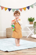 Child in a mustard yellow dress holding a toy in a room with colorful flags and plants.