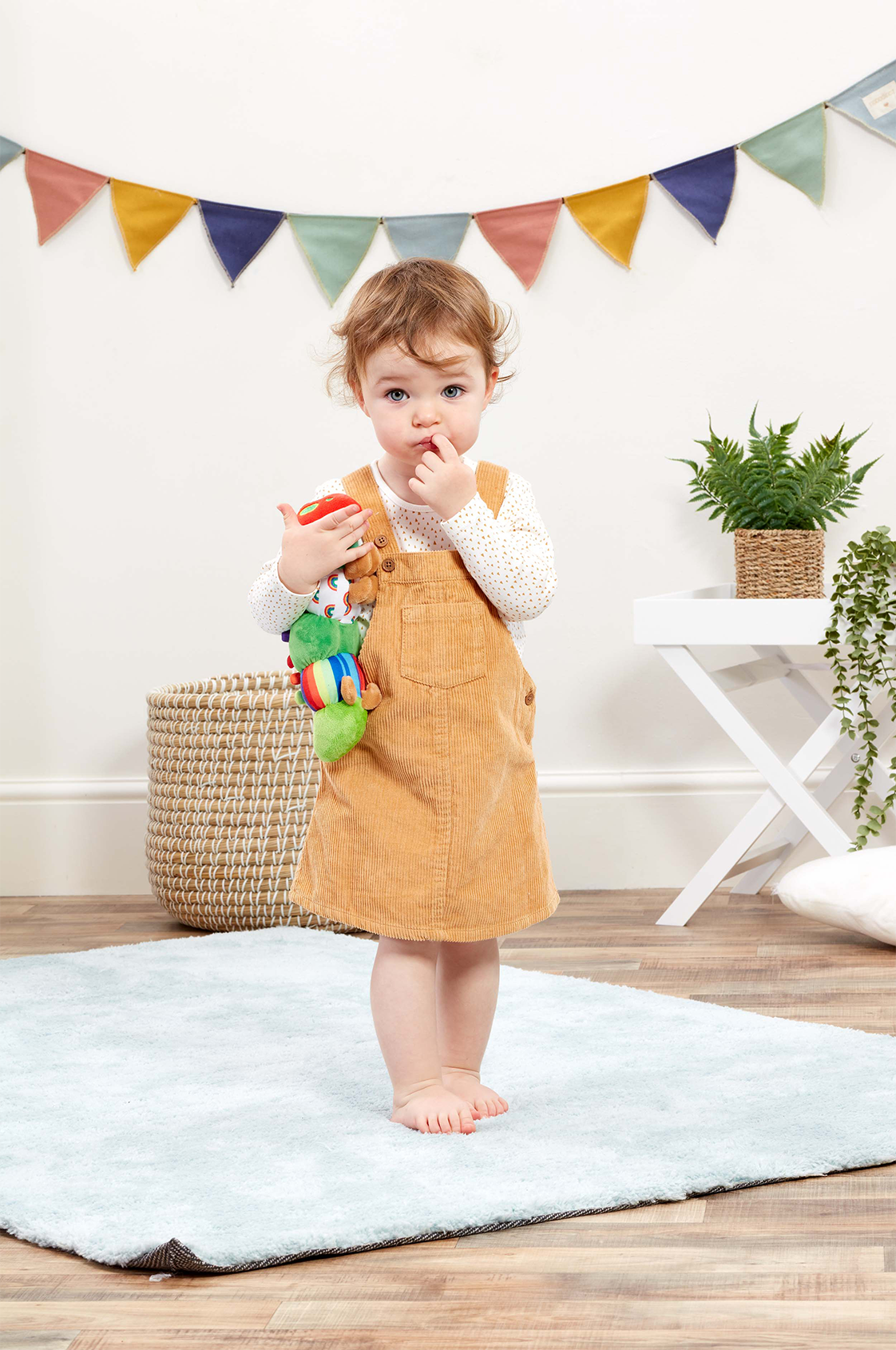 Child in a mustard yellow dress holding a toy in a room with colorful flags and plants.