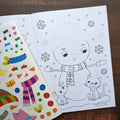 Coloring book with a snowman and cat illustration and a set of related stickers on a wooden surface.