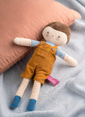 Toy doll with brown hair, orange overalls, and blue shoes lying on a gray surface with a pink pillow.