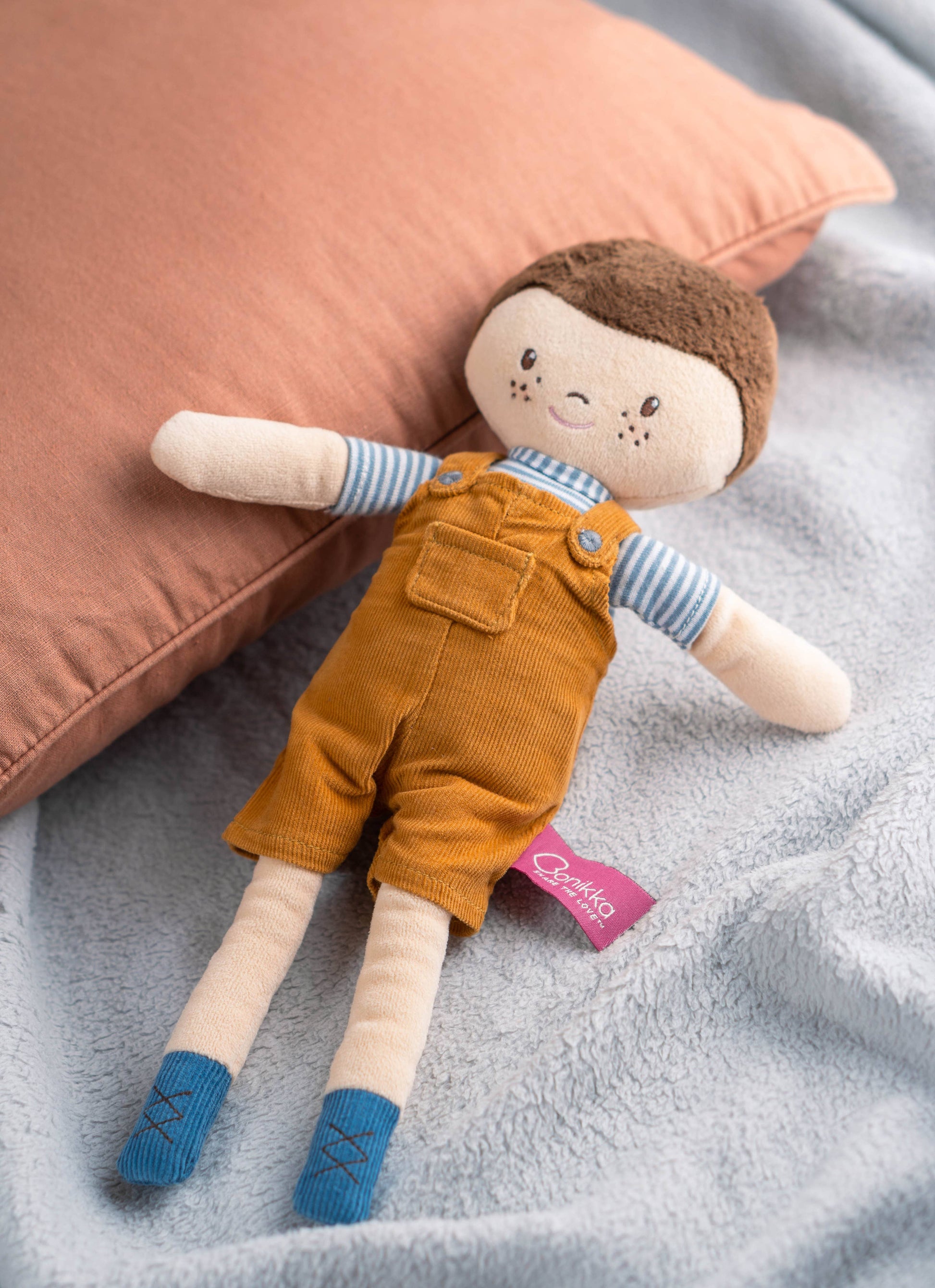 Toy doll with brown hair, orange overalls, and blue shoes lying on a gray surface with a pink pillow.