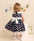 Child wearing a navy blue dress with white polka dots and a bow, standing against a beige background with decorative elements.
