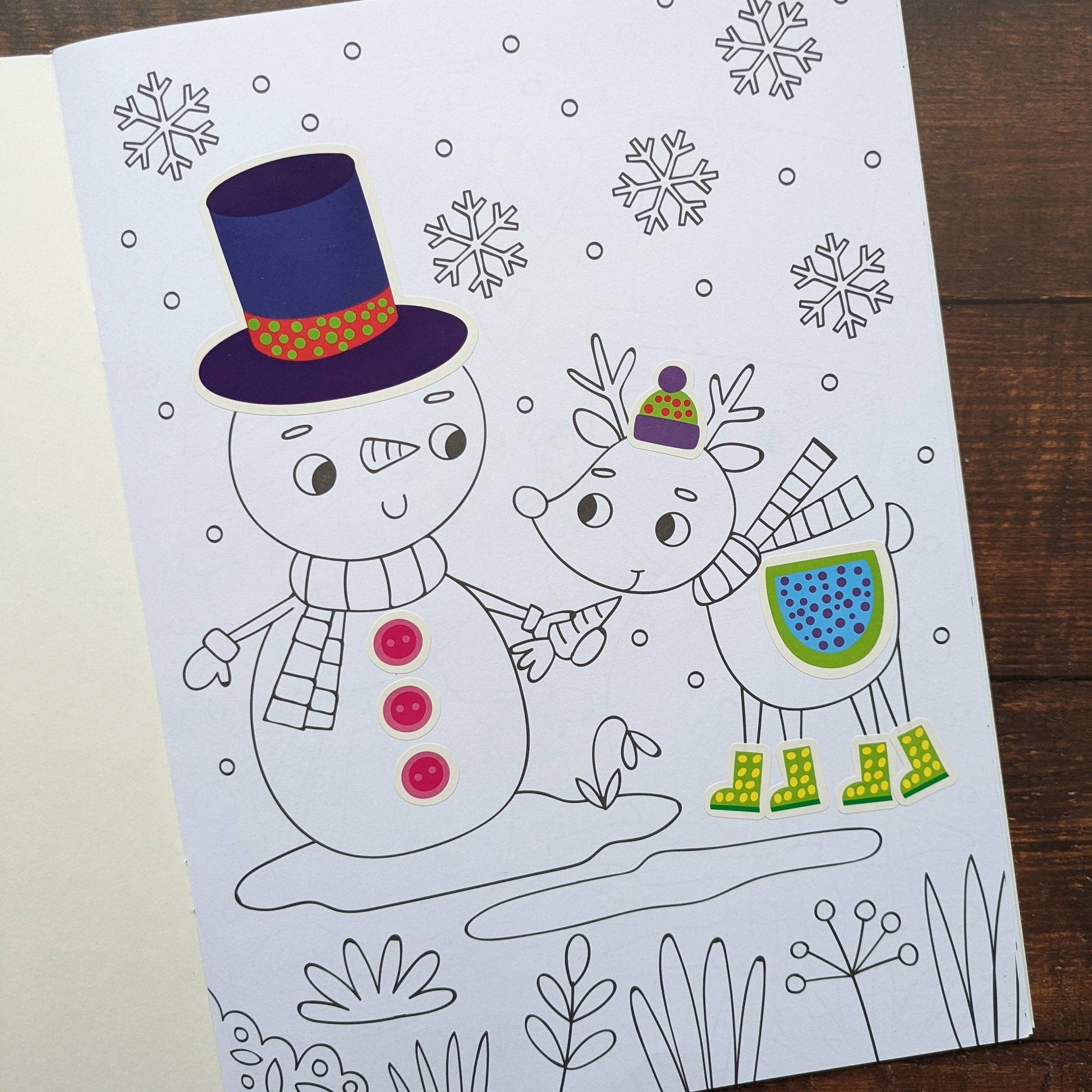Coloring book page with a snowman and reindeer illustration on a wooden surface.