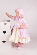 Child in a pink dress and bonnet standing next to a white wooden crate with heart designs.