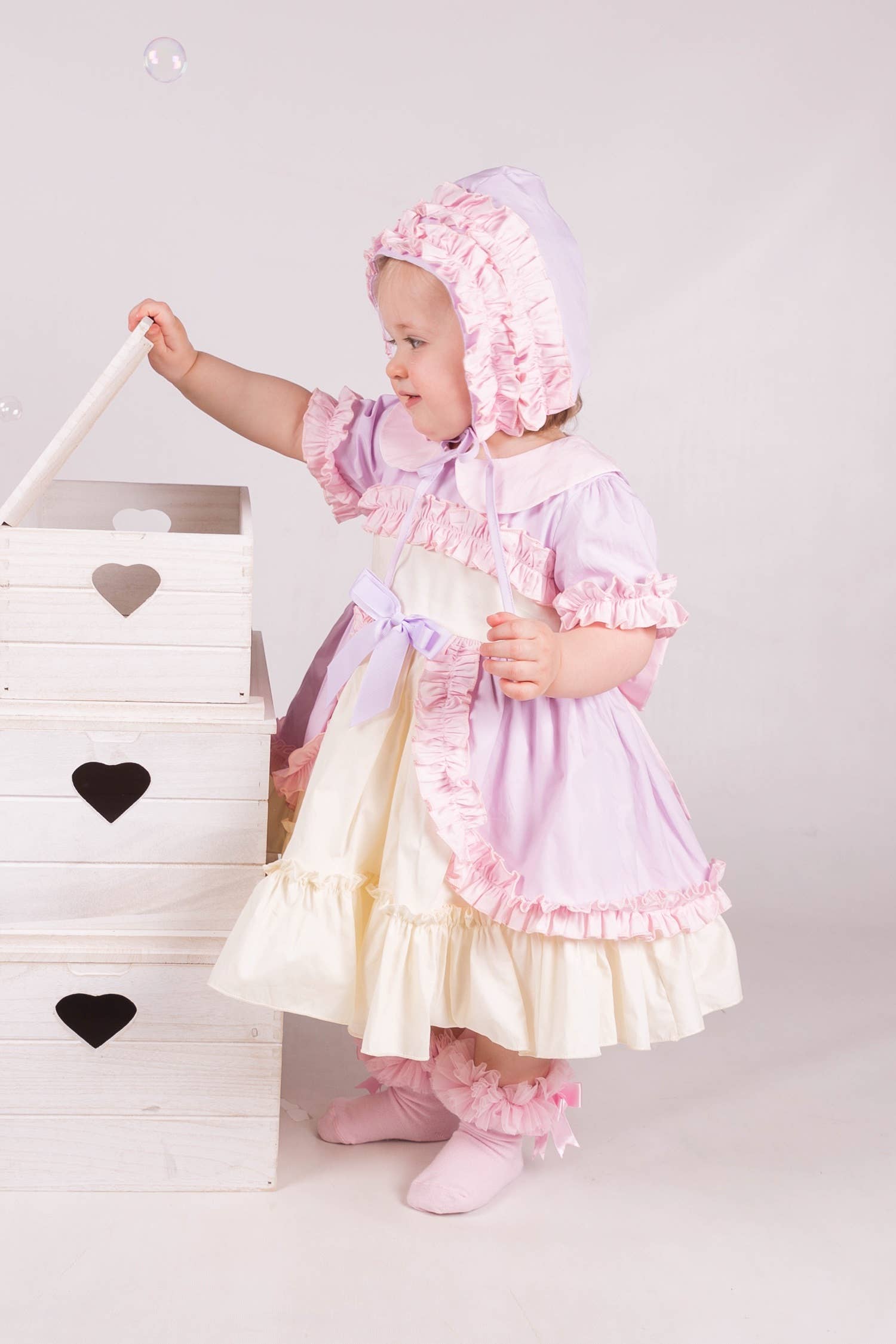 Child in a pink dress and bonnet standing next to a white wooden crate with heart designs.