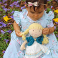 Child in a floral dress holding a doll with braided hair, standing in a garden.