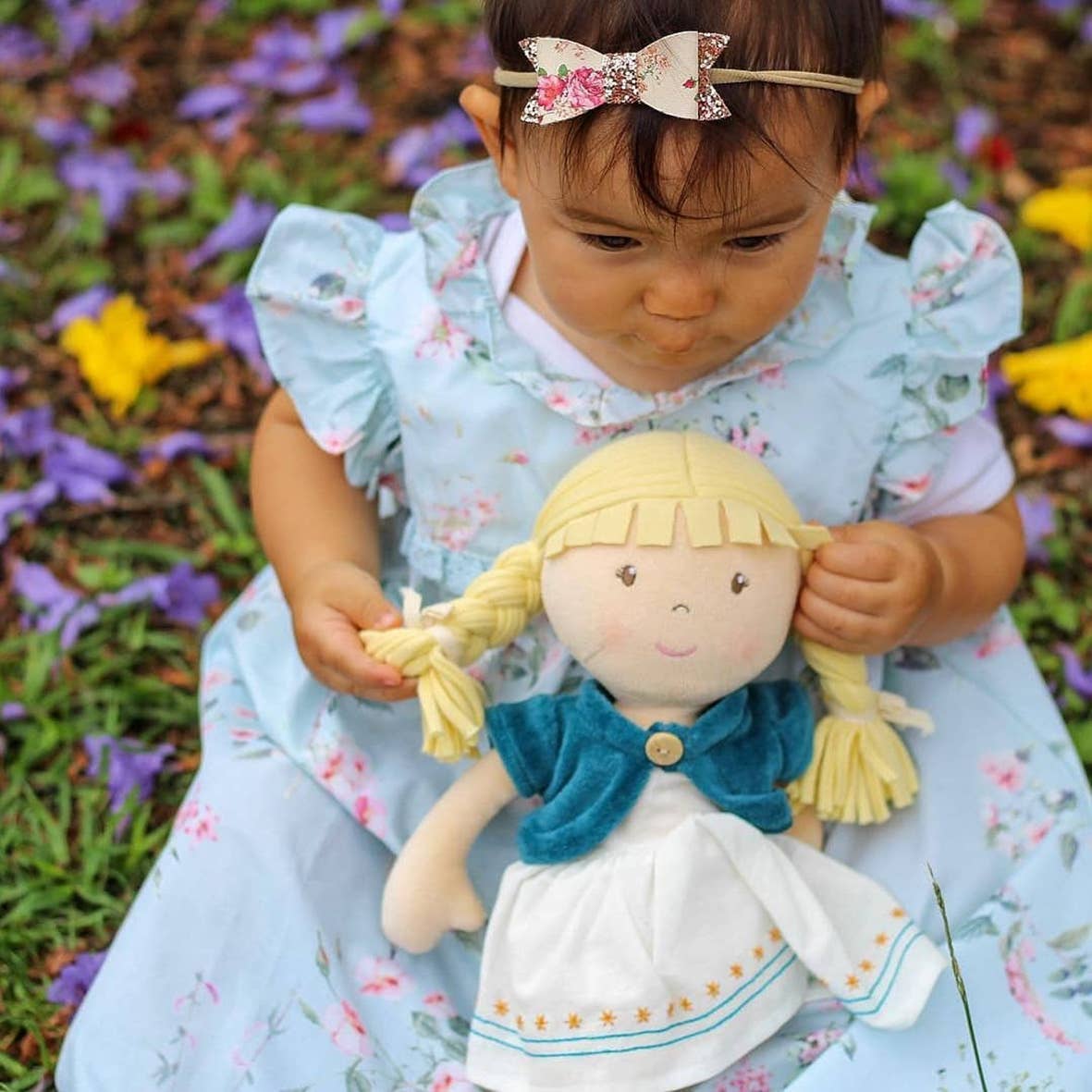 Child in a floral dress holding a doll with braided hair, standing in a garden.
