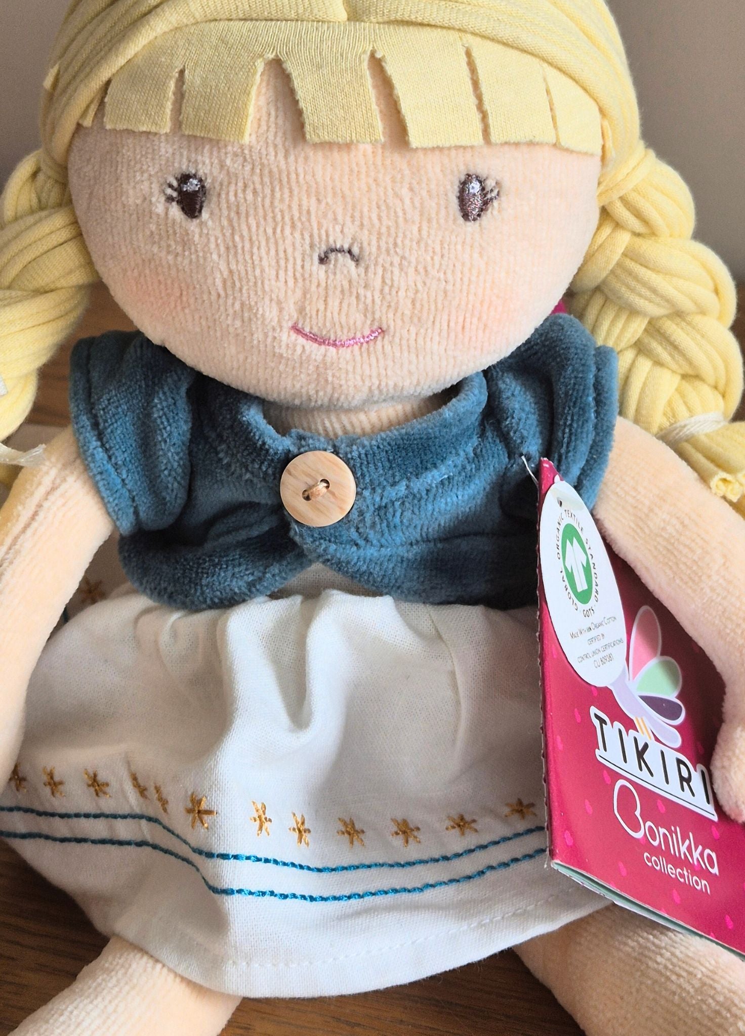 Bonikka organic  ragdoll with blonde hair and blue top, wearing a white dress with embroidery, on a brown background.
