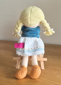 Bonikka ragdoll with blonde hair in plaits wearing a blue jacket and white dress on a wooden surface.