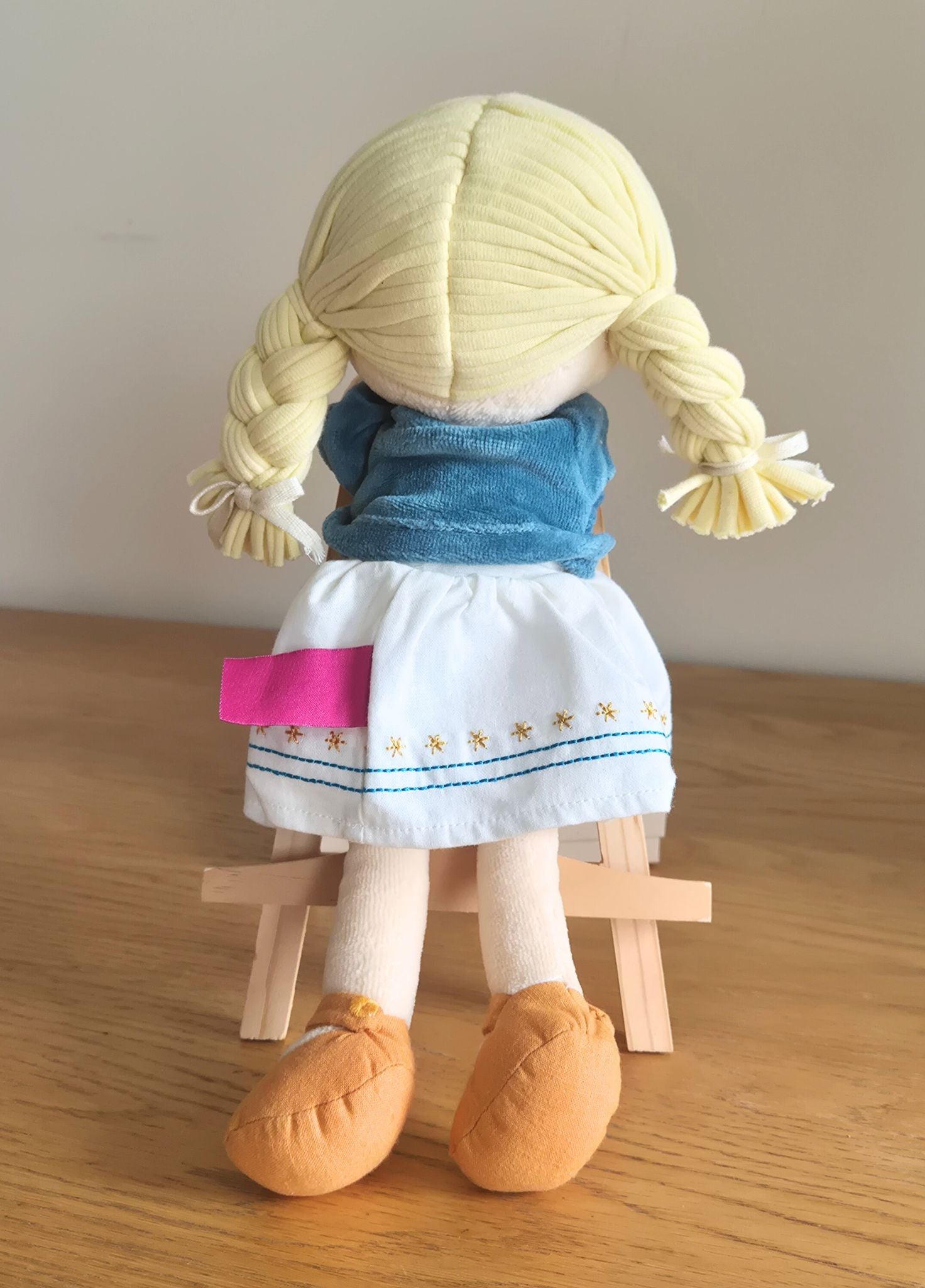 Bonikka ragdoll with blonde hair in plaits wearing a blue jacket and white dress on a wooden surface.