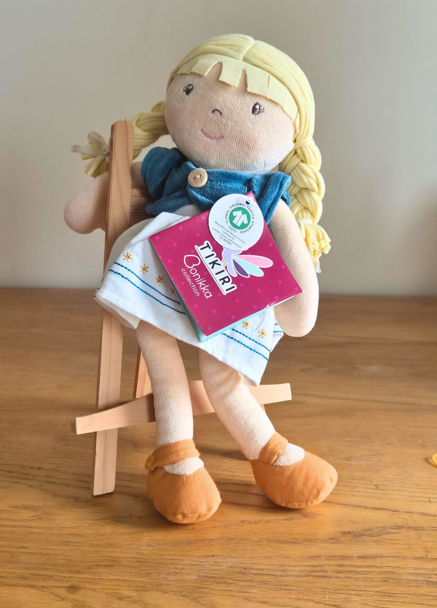 Bonikka organic doll with a blue dress and yellow shoes sitting on a wooden stool, with a visible brand tag.