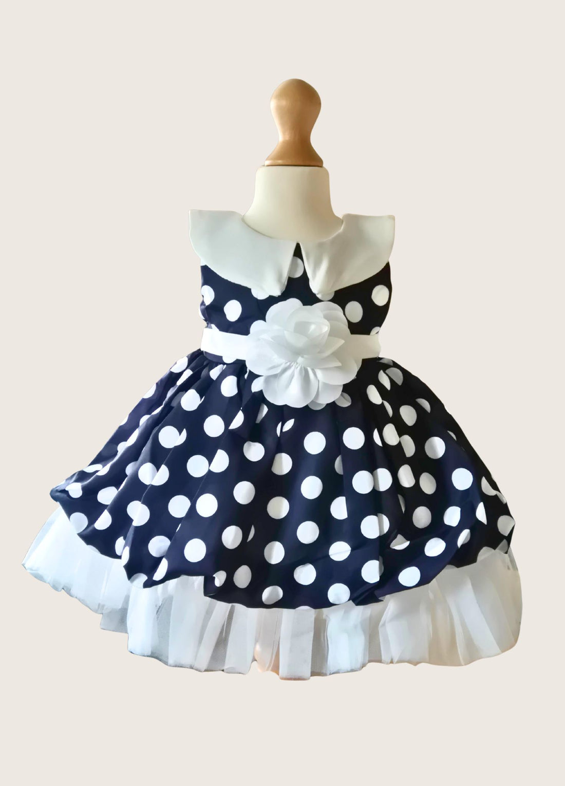 blonde hair girl in a navy and white polka dot dress with white band and white flower at the waistline on a pale background
