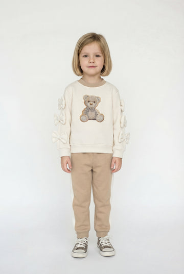 Child wearing a cream sweater with a teddy bear design and beige pants on a white background