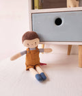 Toy doll with brown hair and overalls sitting on a light wooden floor next to a white dresser.