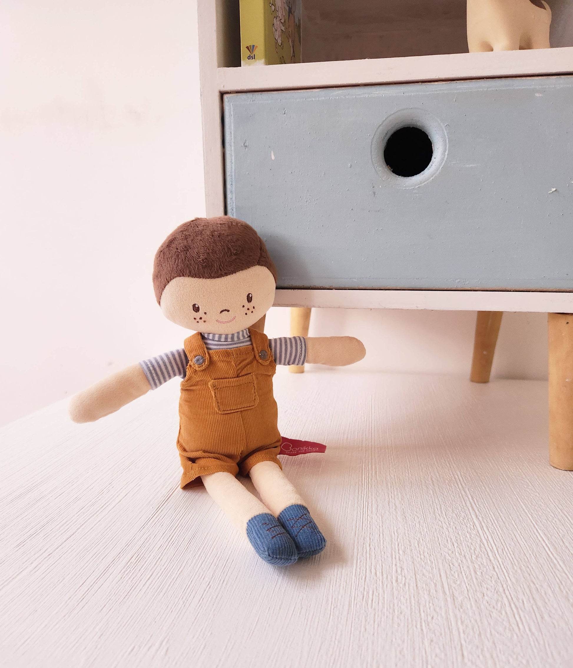 Toy doll with brown hair and overalls sitting on a light wooden floor next to a white dresser.