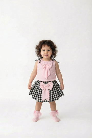 Child wearing a pink top and black and white checkered skirt with a bow on a white background