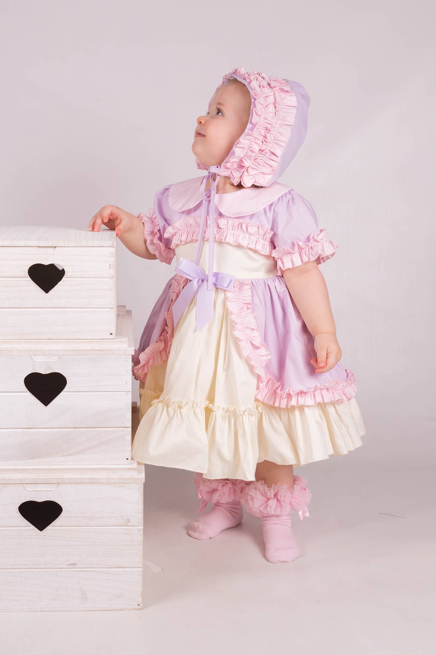 Child in a vintage-style dress and bonnet standing next to a white wooden dresser with black heart designs.