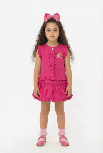 Young girl wearing a pink matching bubble skirt and top with tie detail with a bow on her head against a white background
