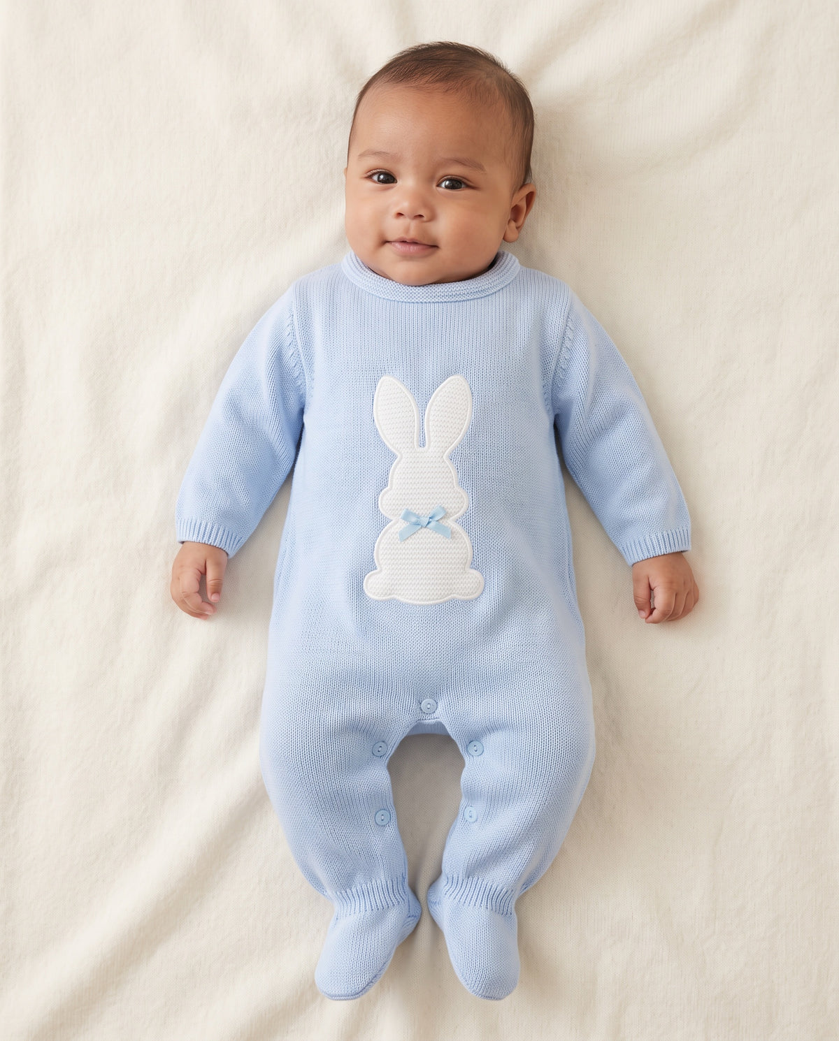 A baby lying on a cream-colored blanket, wearing a light blue ribbed knit footed sleeper with a white bunny applique on the chest.