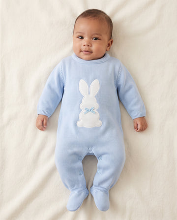 A baby lying on a cream-colored blanket, wearing a light blue ribbed knit footed sleeper with a white bunny applique on the chest.