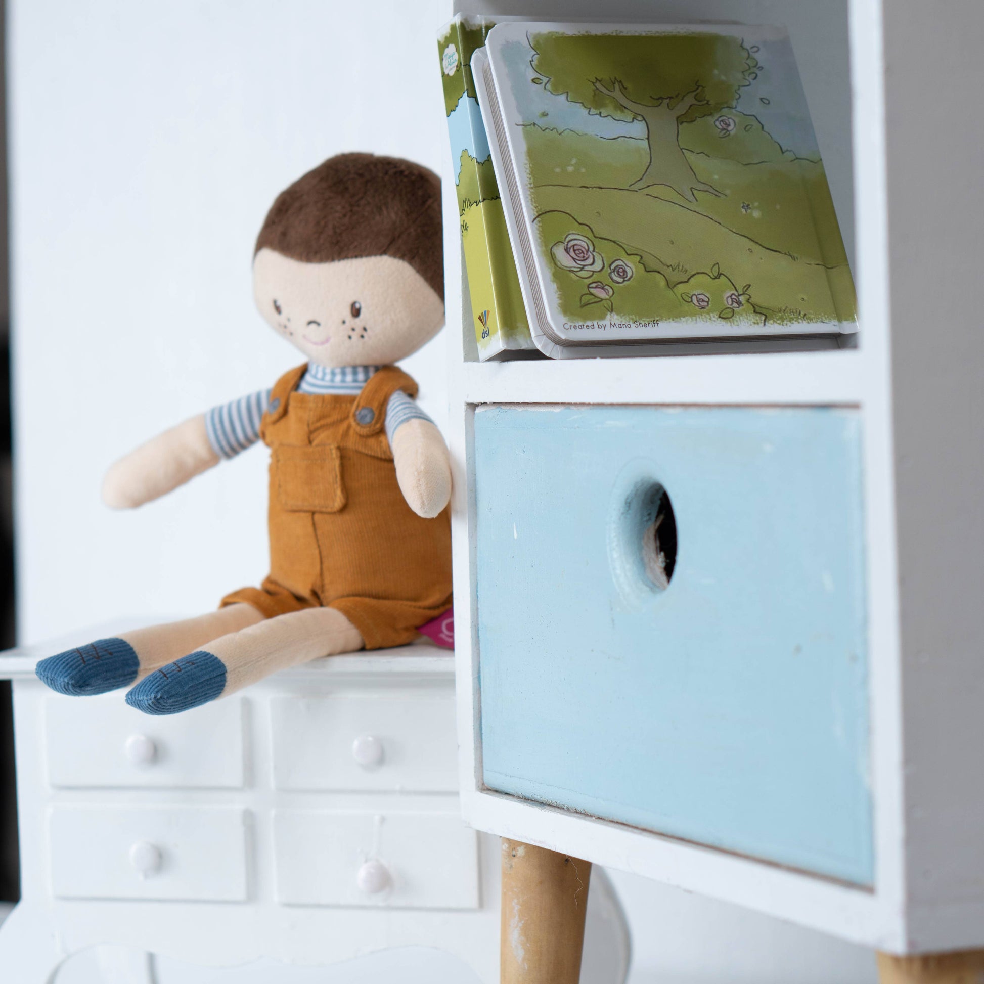 Stuffed doll in overalls next to children's books on a white shelf.