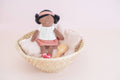Small doll with braided hair sitting in a woven basket on a light pink background