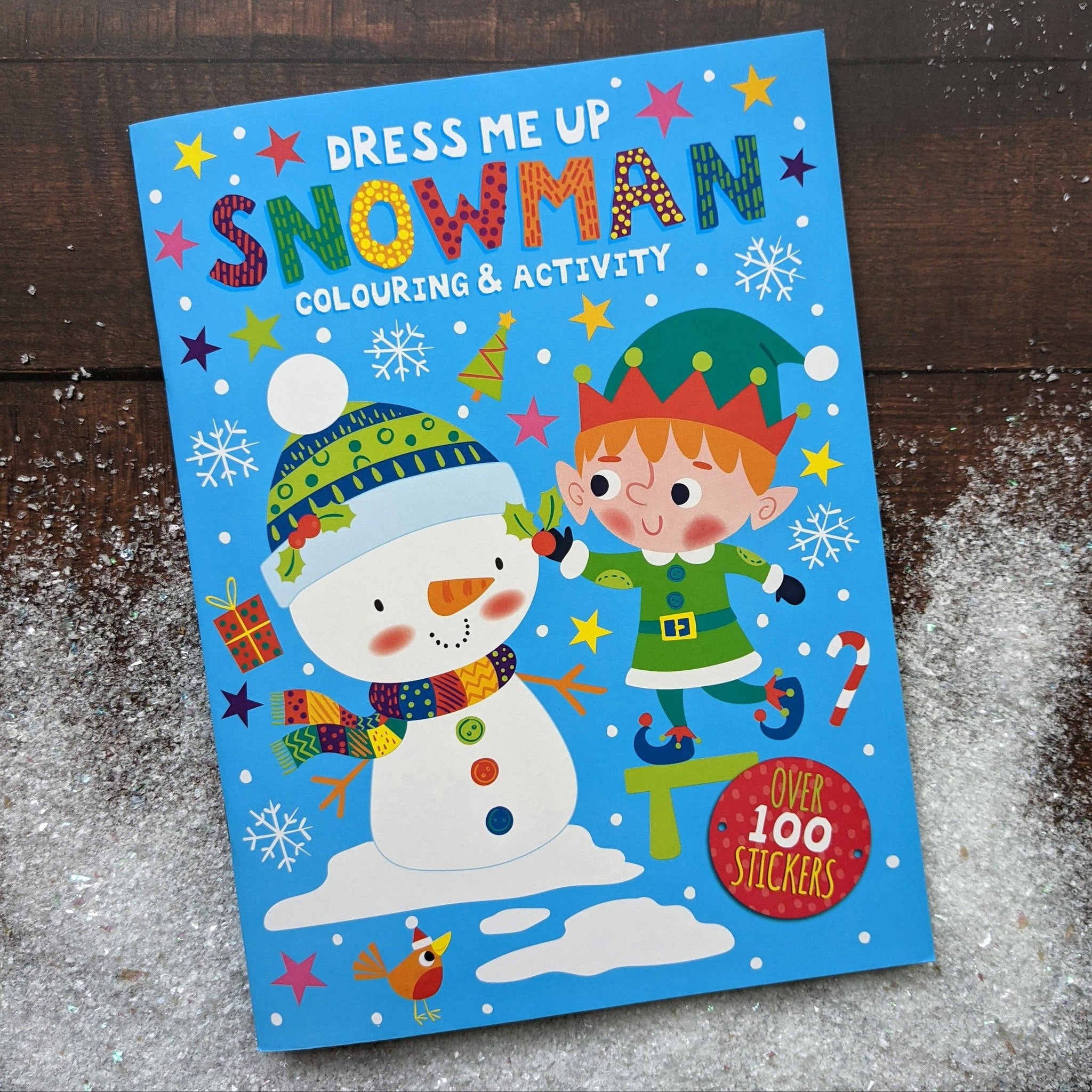 Children's activity book titled 'Dress Me Up Snowman' with a snowman and elf illustration on a wooden surface.