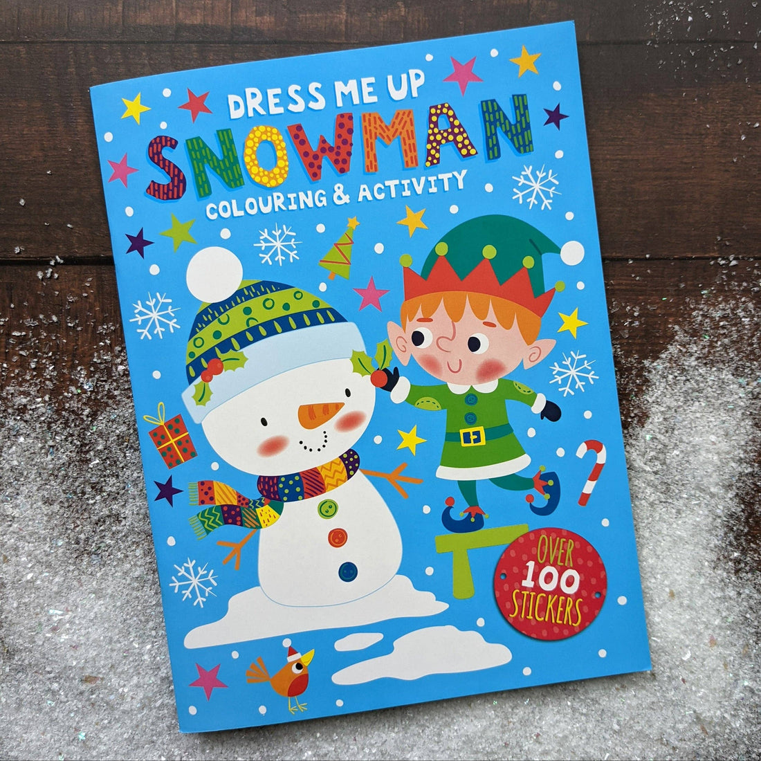 Children's activity book titled 'Dress Me Up Snowman' with a snowman and elf illustration on a wooden surface.