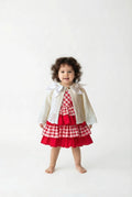 Little girl with dark curly hair wearing a red and white checked dress with an ivory double bow cardigan on top.