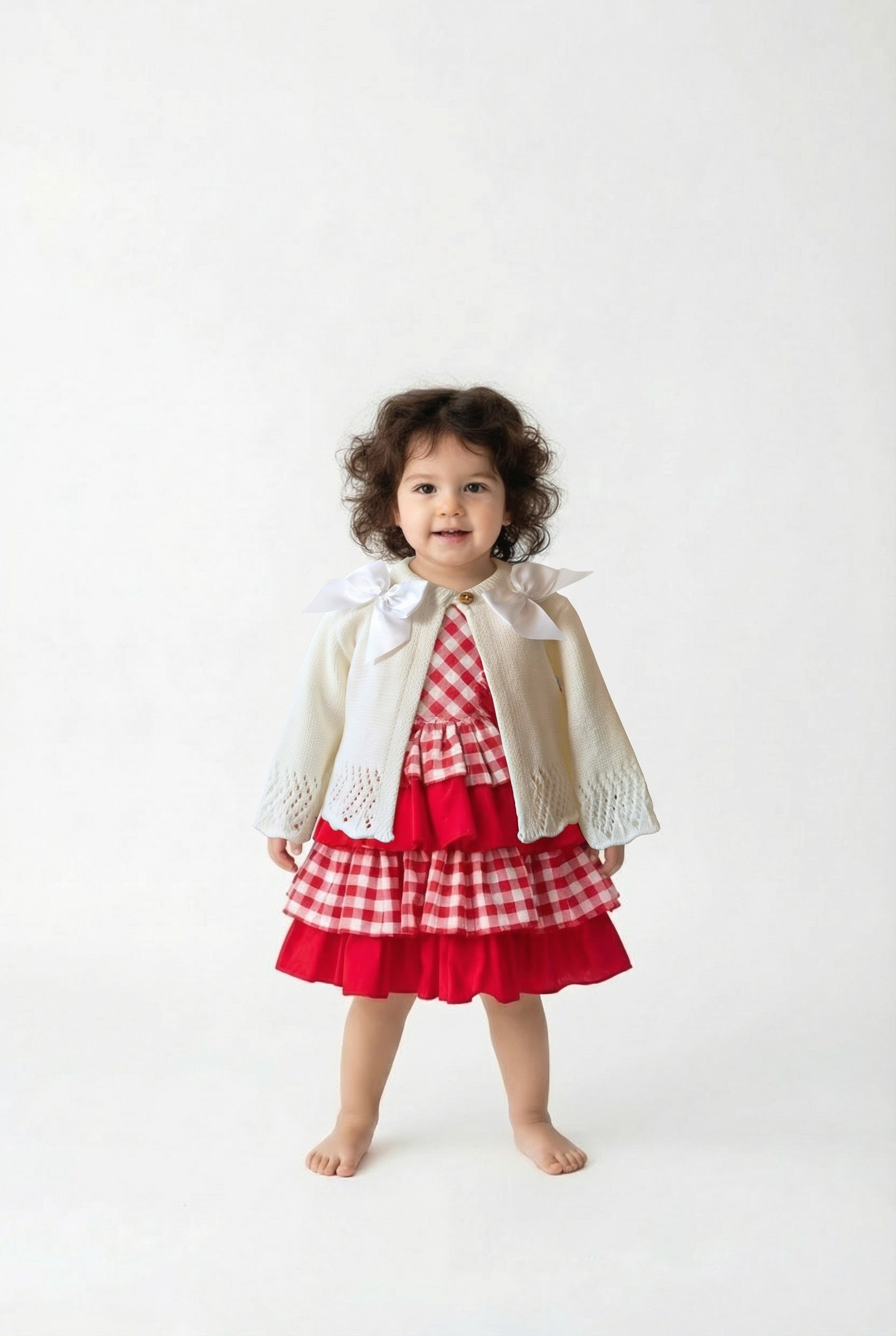 Little girl with dark curly hair wearing a red and white checked dress with an ivory double bow cardigan on top.