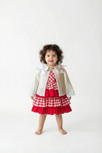Little girl with dark curly hair wearing a red and white checked dress with an ivory double bow cardigan on top.
