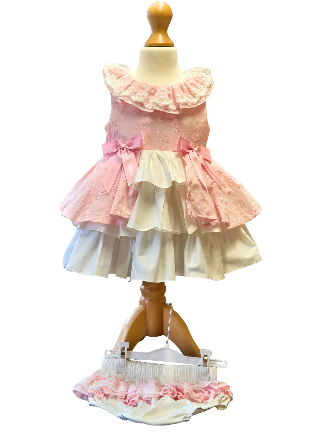 Toddler with curly black hair in a pink and white tiered peplum dress with pink bows on a pale background