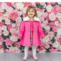 Child wearing a pink coat with white fur trim in front of a floral wall.
