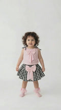toddler in a black and white gingham skirt with pink bow and a pink top with gingham shoulder ties on a light background