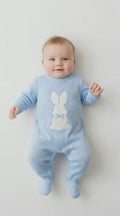 baby boy in a blue all in one knitted romper with a white rabbit in the centre on a light coloured background.
