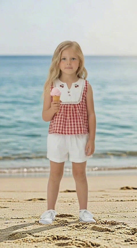 Blonde haired girl standing on a beach wearing a red and white gingham sleeveless top with embroidered cherries near the neckline and ivoru shorts.