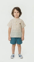 young boy aged around 4 in a neutral linen shirt with petrol blue large checks, a grandad collar and co ordinating petrol blue shorts on a white background.