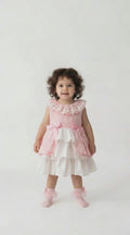 Toddler with curly black hair in a pink and white tiered peplum dress with pink bows on a pale background