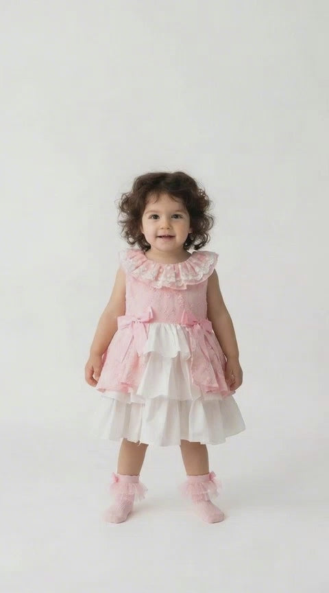Toddler with curly black hair in a pink and white tiered peplum dress with pink bows on a pale background