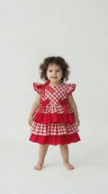 Video of a toddler with black curly hair in a red and white gingham check tiered dress.  Smiling and swaying her hips.