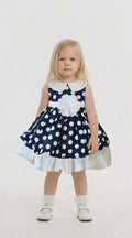 blonde hair girl in a navy and white polka dot dress with white band and white flower at the waistline on a pale background