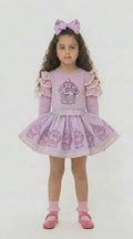 Young girl in a lilac frilled top with a sparkly cupcake design and a co ordinating lilac skirt that has cupcakes around the hemline.