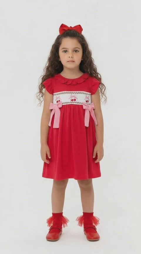Young girl in a red dress with smocking and embroidered cherries and pink material bows on a light background.