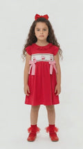 Young girl in a red dress with smocking and embroidered cherries and pink material bows on a light background.