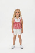 Young girl wearing a red and white gingham top with a white bibbed neckline and ivory shorts on a white background.