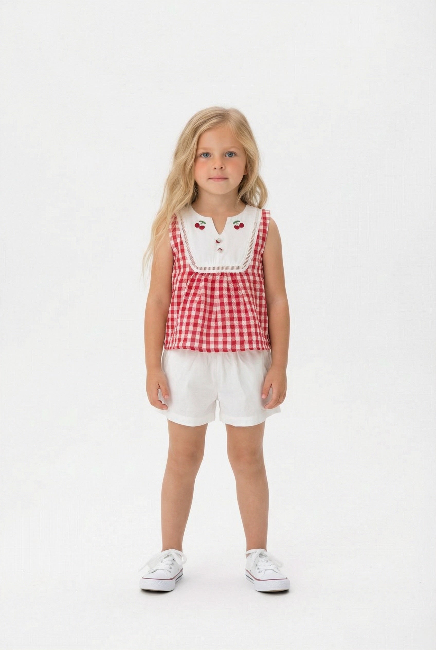 Young girl wearing a red and white gingham top with a white bibbed neckline and ivory shorts on a white background.