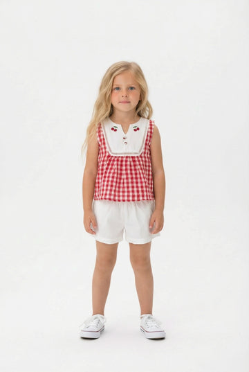 Young girl wearing a red and white gingham top with a white bibbed neckline and ivory shorts on a white background.